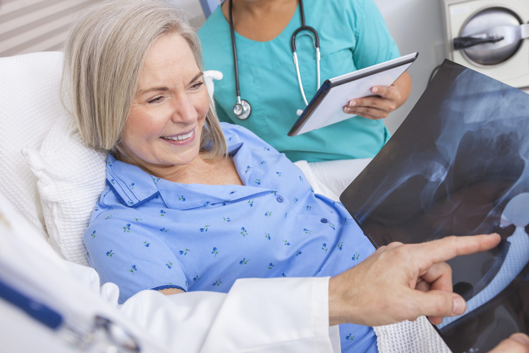  A doctor explains a hip X-ray to a patient in a hospital bed with a nurse standing by. 