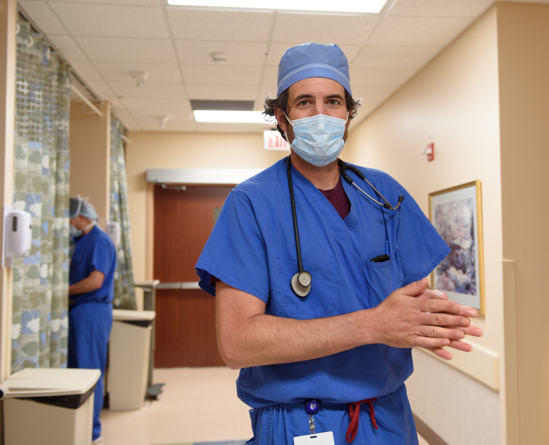 A plastic surgeon stands in OR corridor, dressed in blue scrubs and mask, getting ready for breast reduction surgery. 