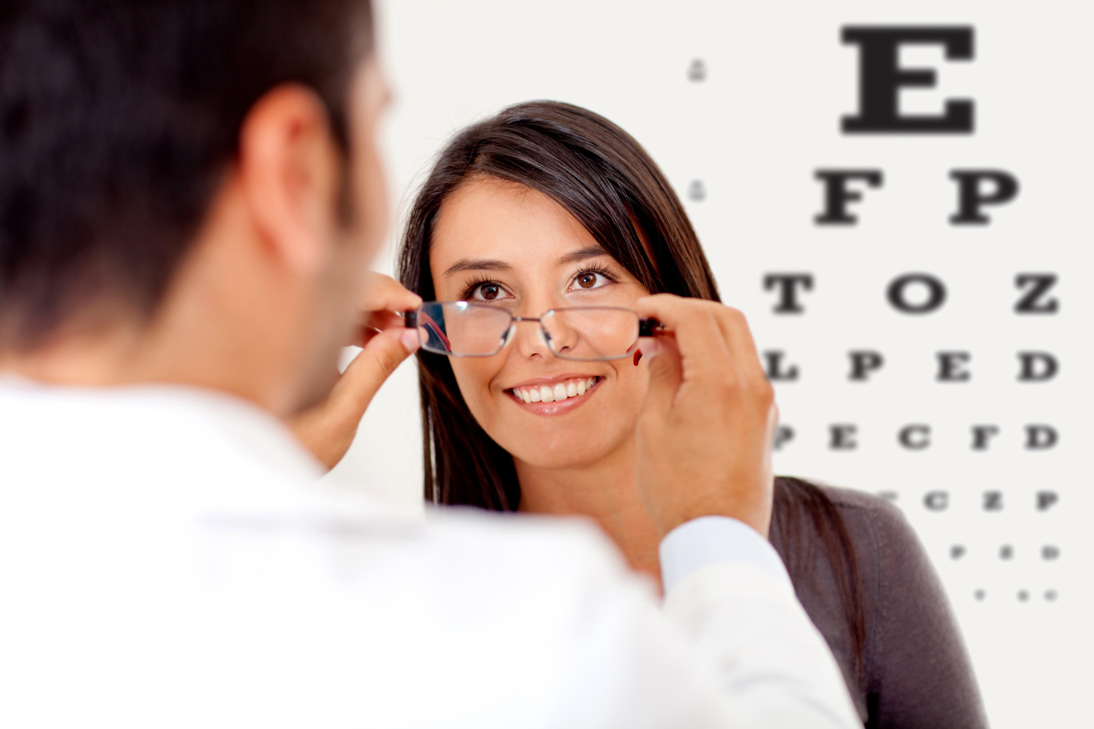 A male eye doctor helps a young woman standing in front of an eye exam chart choose glasses to help reduce eye strain. 