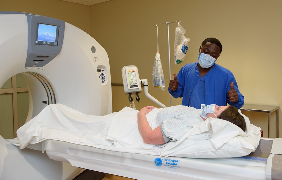 A staff member in scrubs and a mask assists a patient who is having a CT scan.