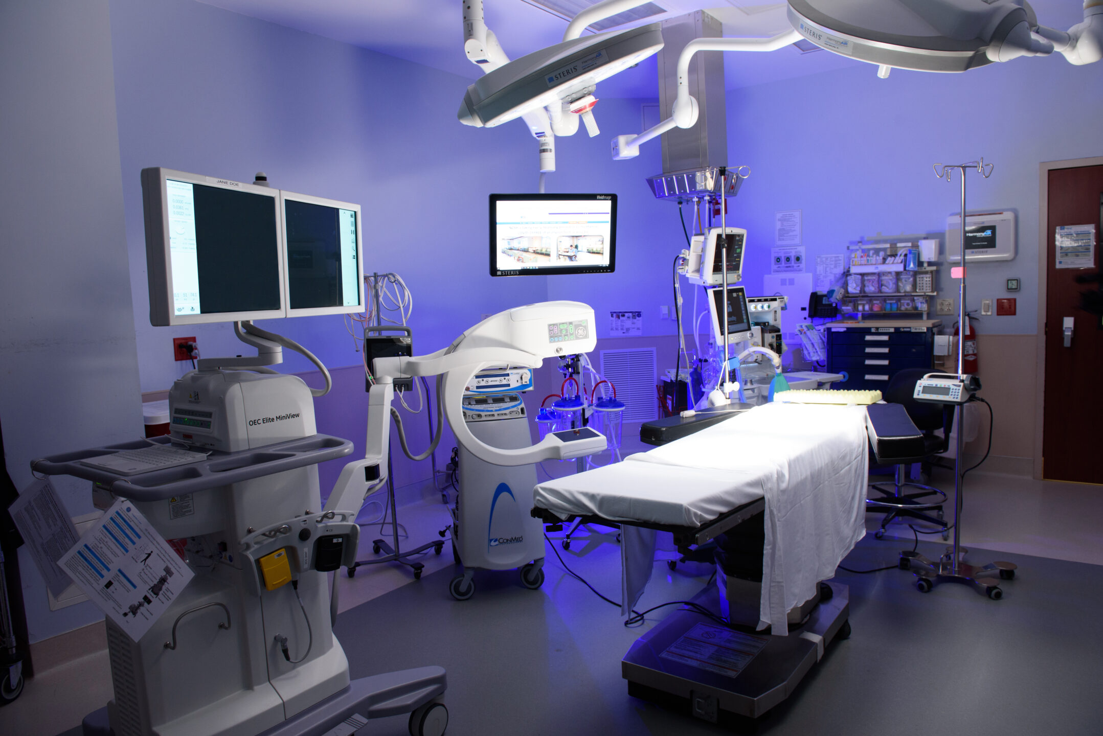 An image of the operating room is lit with blue light and shows a bed and surgical equipment.