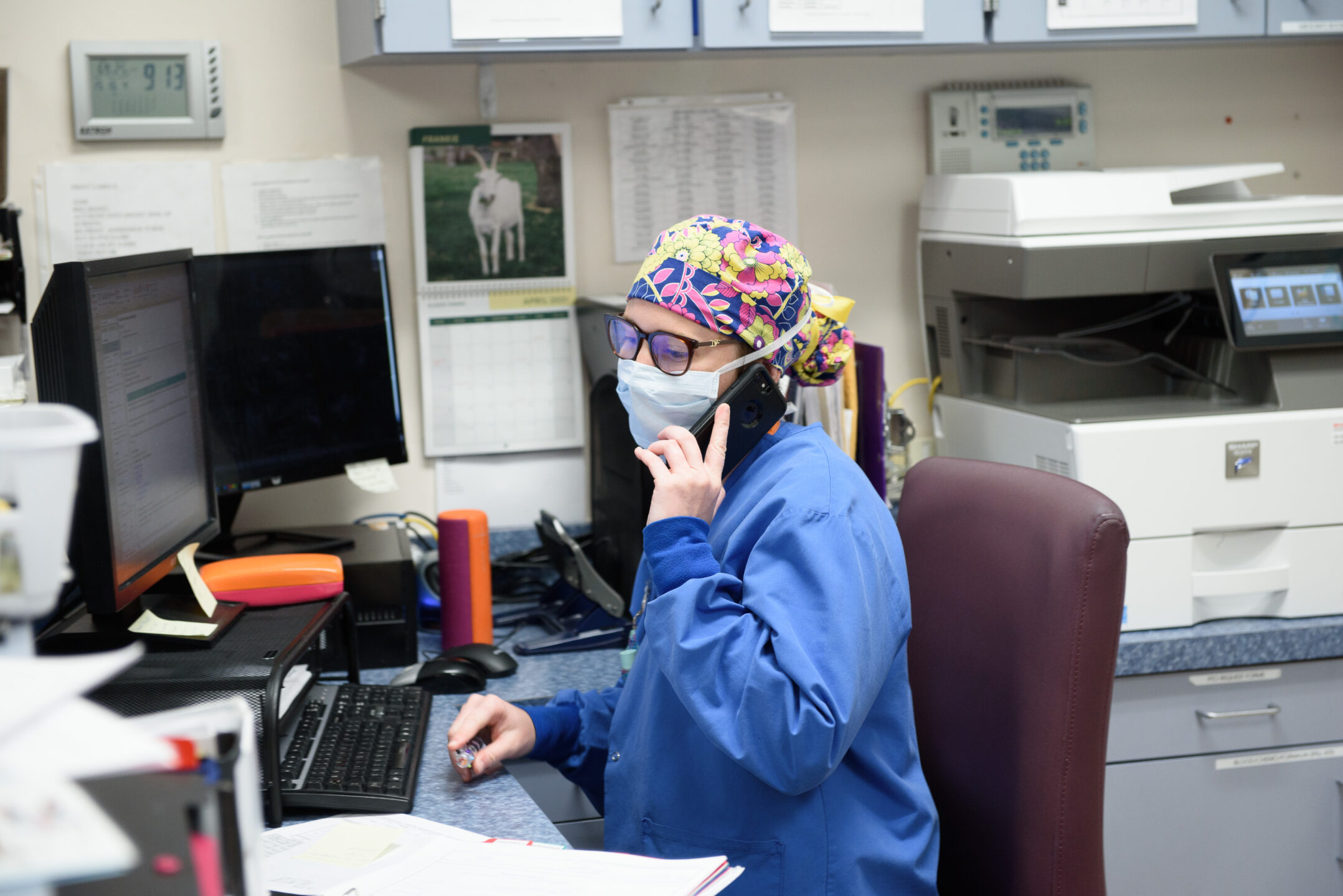 A female staff member in scrubs and a mask talks on the phone behind a desk and computer. 