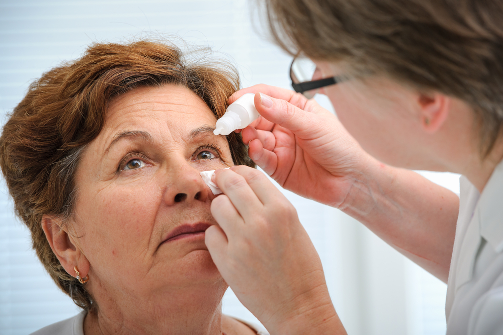 An older woman leans back as an eye doctor puts drops in her eye because she has eye pain from blinking. 
