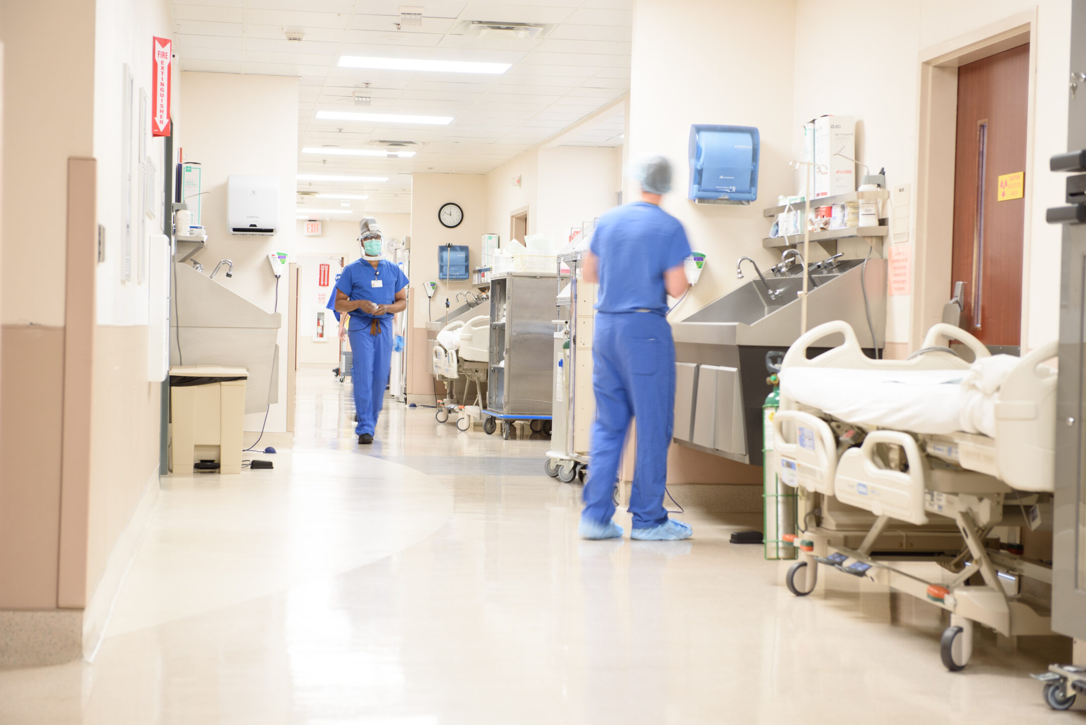 A hospital hallway shows a staff member washing his hands and another walking toward him.