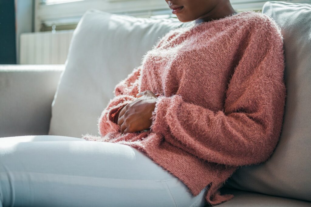 A woman sits on a couch, holding her stomach in pain.