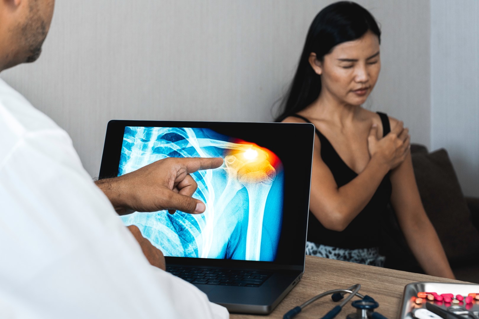A woman holds her hand to her sore shoulder while the doctor looks at an image of her rotator cuff tear on a screen.