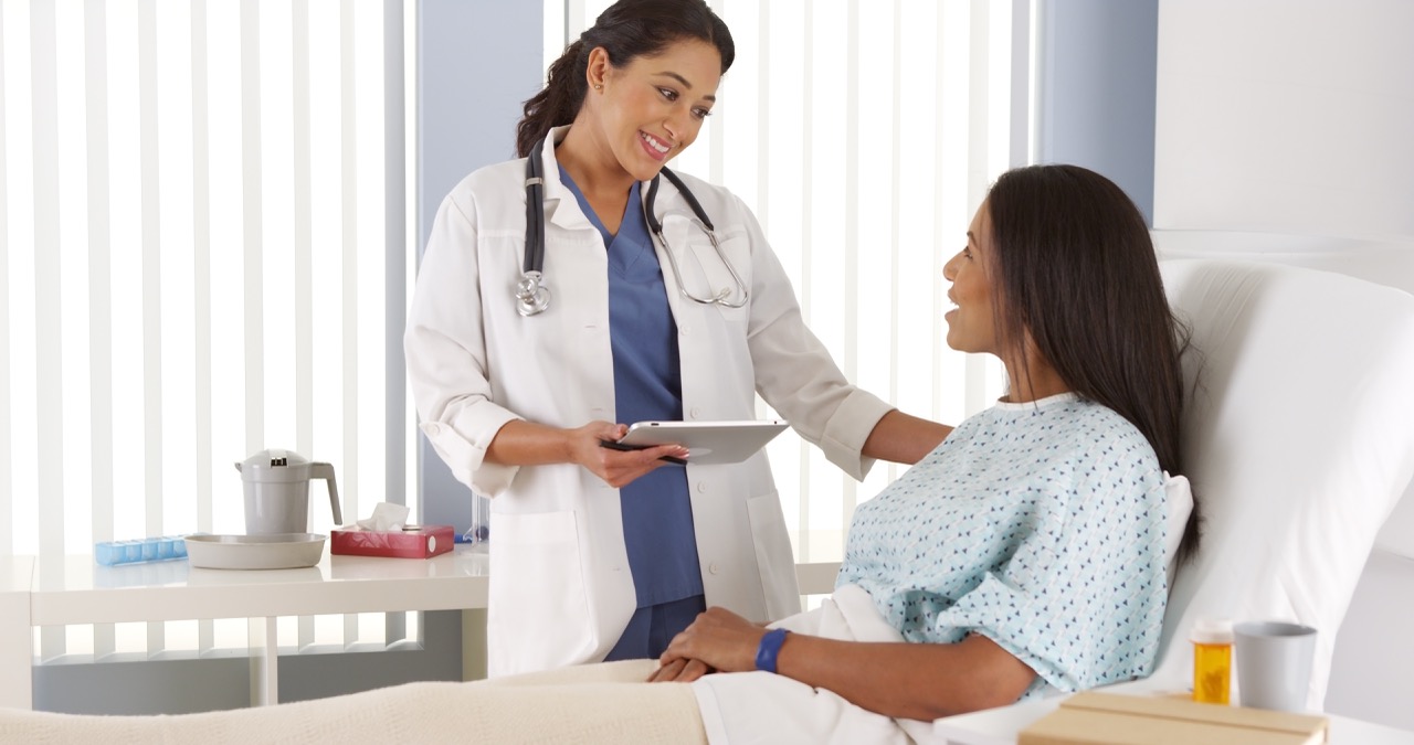 A doctor speaks to a patient dressed in a hospital gown and sitting on a hospital bed.